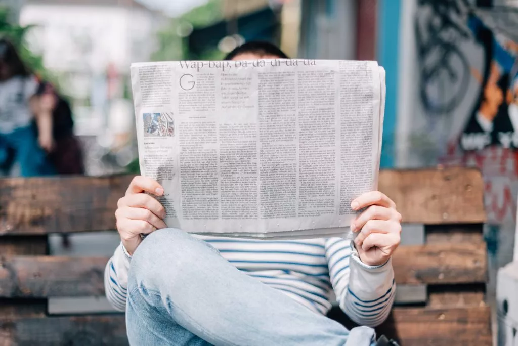 Person in blue and white striped shirt sitting on park bench reading the newspaper